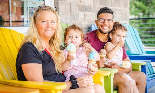 A family sitting in colourful Adirondack chairs eating ice cream. There is a grandma, a dad, and two children.